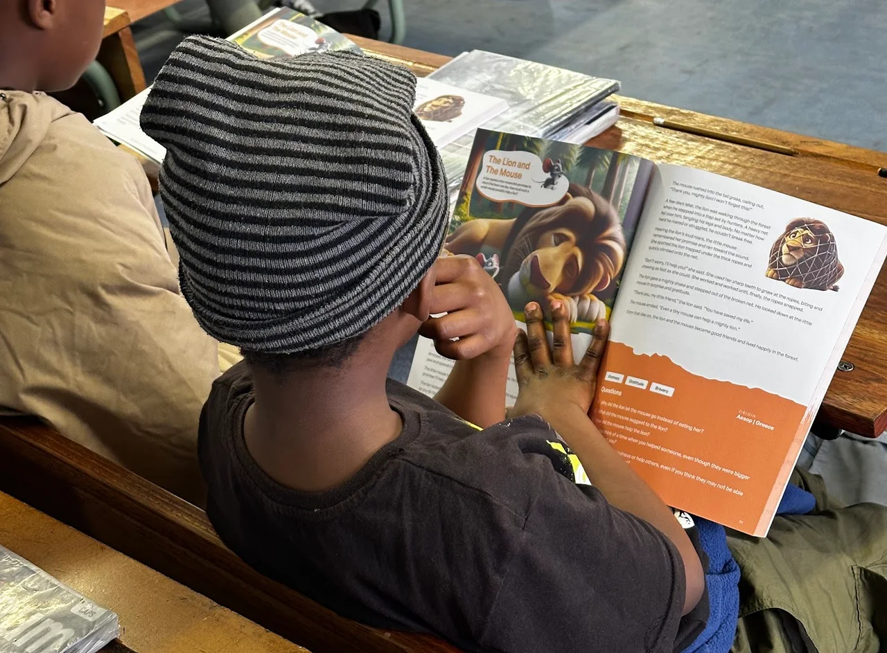 A learner seen from behind reading The Lion and the Mouse in a FableReads book at a classroom desk.