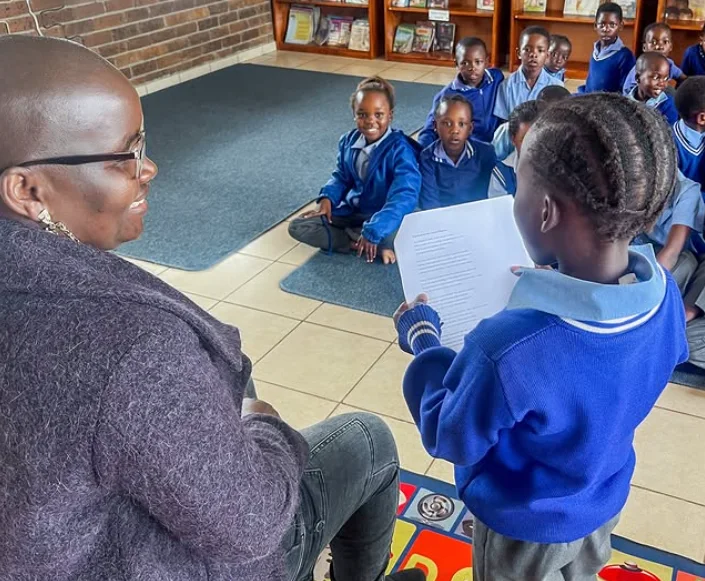 A learner reads a story aloud to classmates and a facilitator in a Kaross Foundation literacy session.