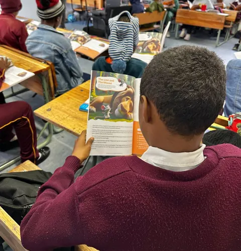 Ein Kind liest, von hinten gesehen, „Der Löwe und die Maus“ in einem FableReads-Buch an einem Schultisch.